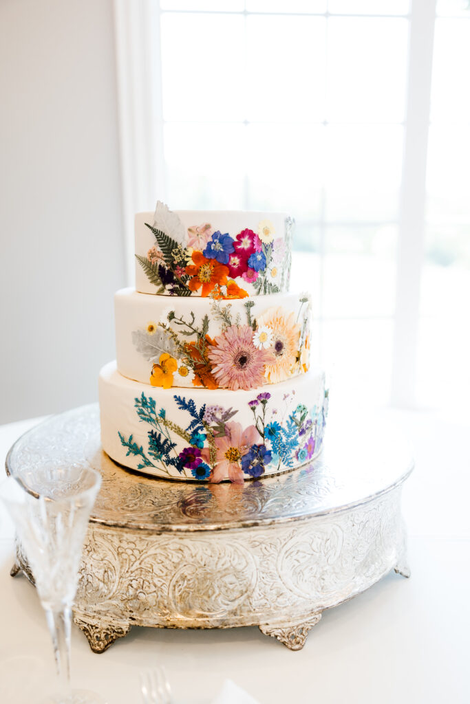 a watercolor floral wedding cake sits on a antique silver cake stand at the wedding reception held at milestone mansion in denton, texas.