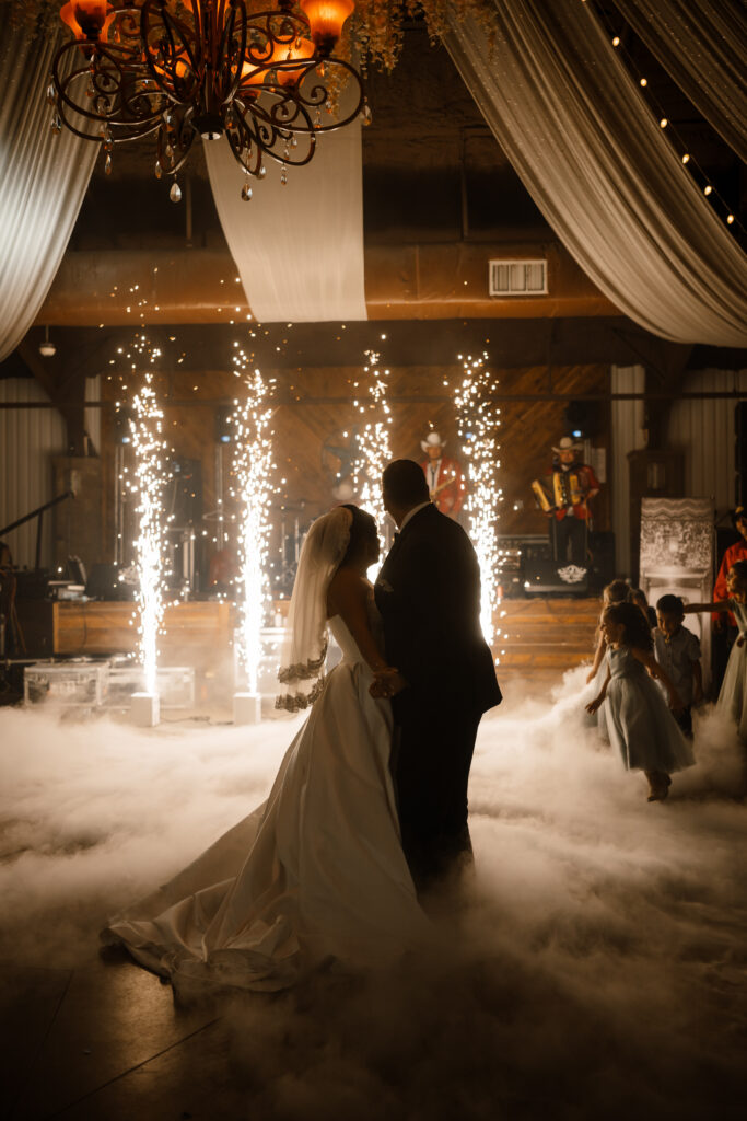 the bride and groom hare their first dance as cold sparks blast behind them and fog wraps them up while children dance around them.
