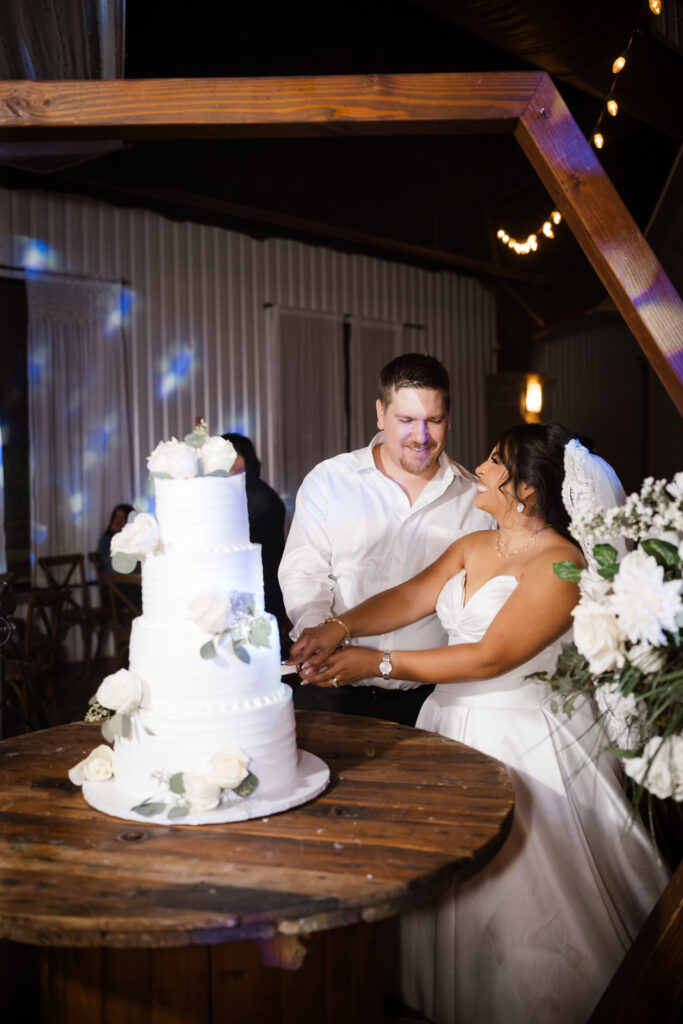 The bride and groom share a smile as a they slice their 3 tiered wedding cake while Shelly Voss document the moment.

