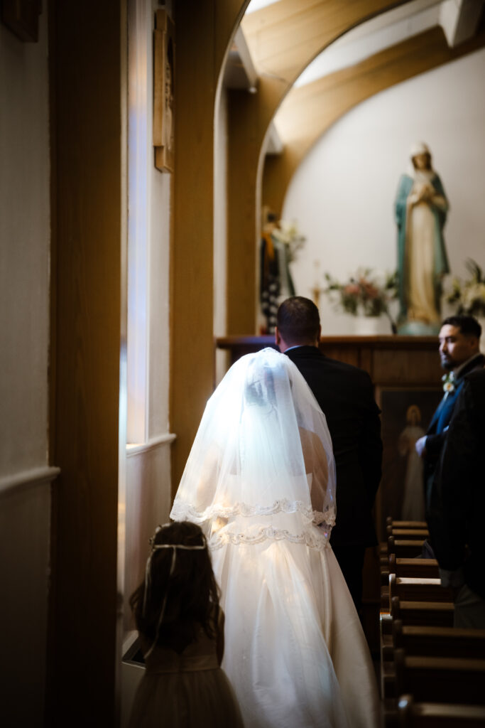 A bride walks by a stained glass window and a rainbow is reflected in her vail at the catholic church
