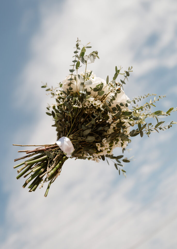 the modern day shot of the bouquet flying through the air with a blue sky and cloud in the background captured by shelly voss
