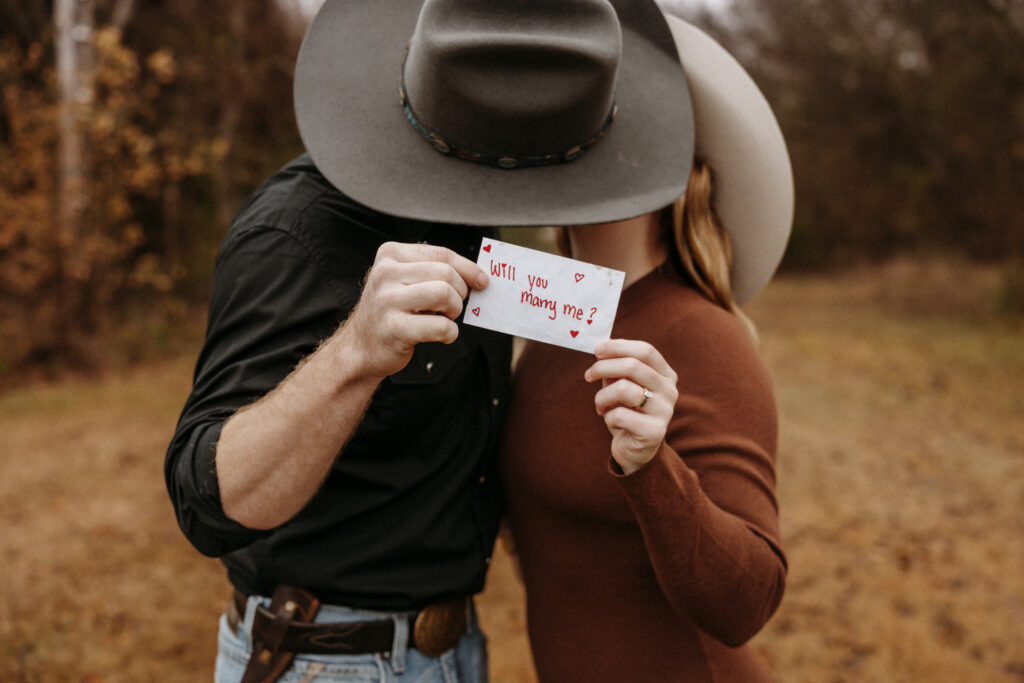 couple shares a kiss holding a note asking her to marry him while Shelly Voss photography captures the moment